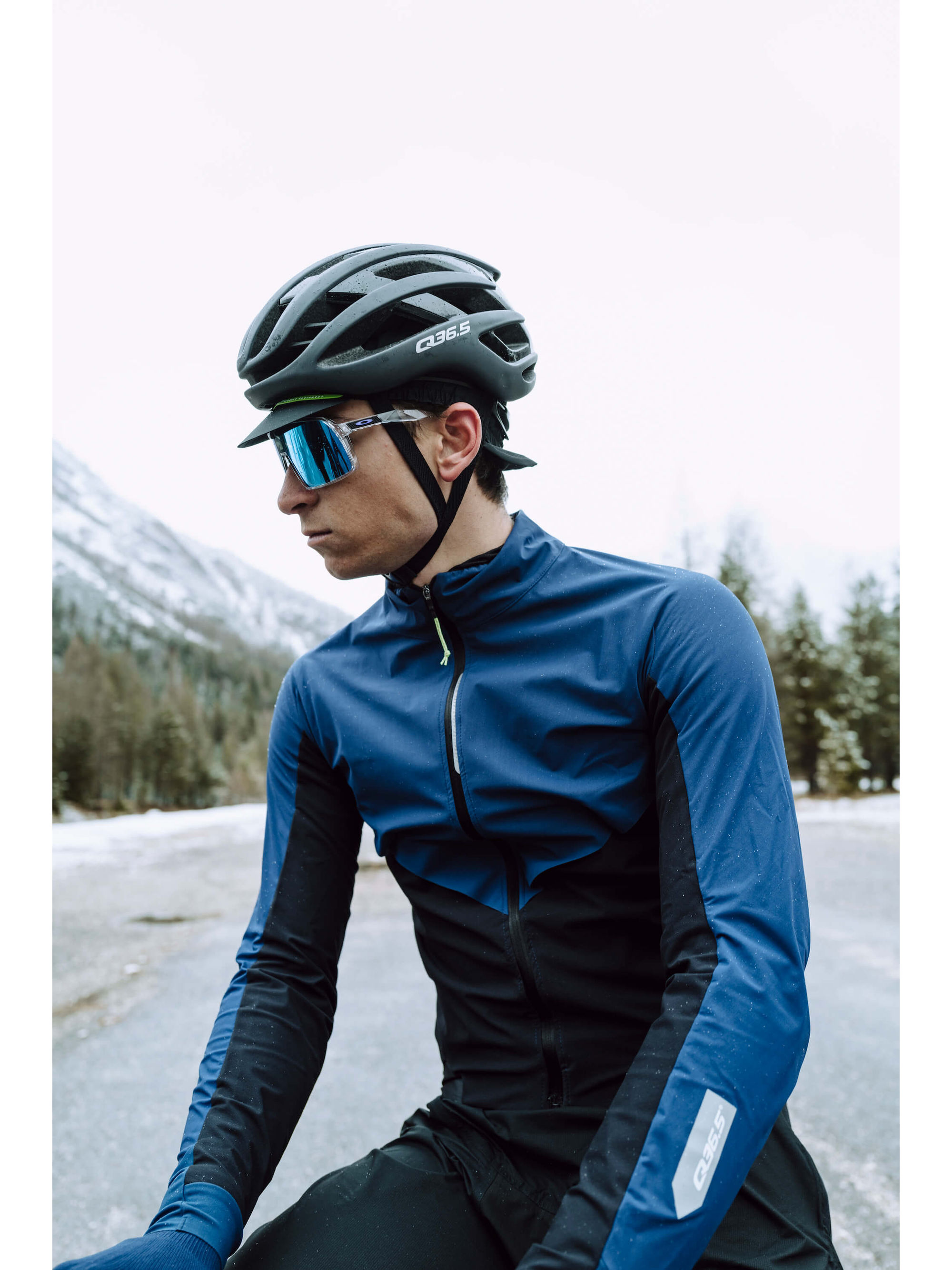 A cyclist wearing the blue Q36.5 Bat Shell Long Sleeve Jersey, demonstrating its fit and temperature control features in a mountainous setting.