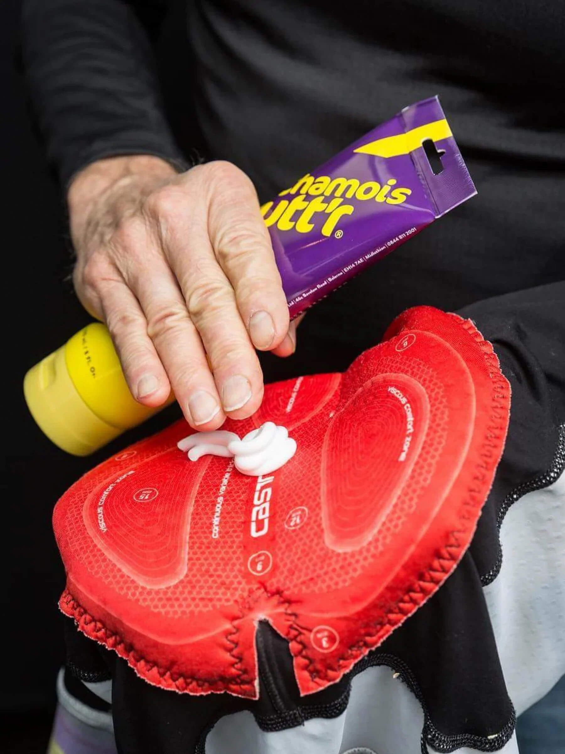 An image of an 8oz bottle of Unisex Chamois cream by Chamois Butt'r being squeezed onto a cycling bib chamois, this is a premium chamois cream for any cyclist.