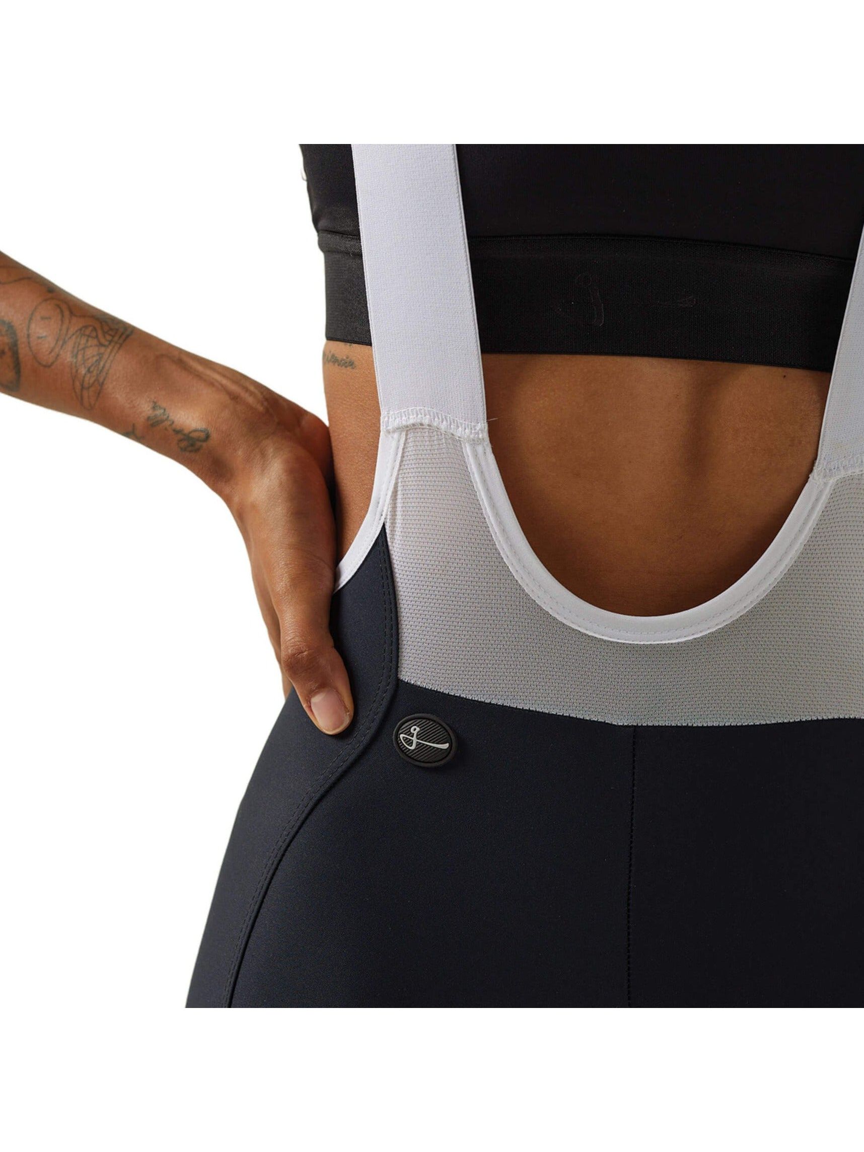 Close-up side view of a woman's lower torso wearing black Givelo HD Pro Bib Shorts, showing the detailed stitching and fabric texture, along with the white shoulder straps and the brand's button logo near the hip.