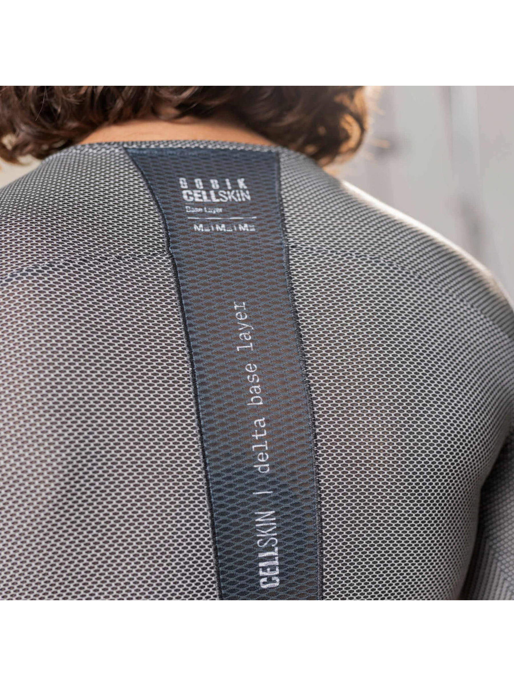 The back view of a man wearing a grey, short-sleeved base layer cycling shirt. A central black stripe with the text 
