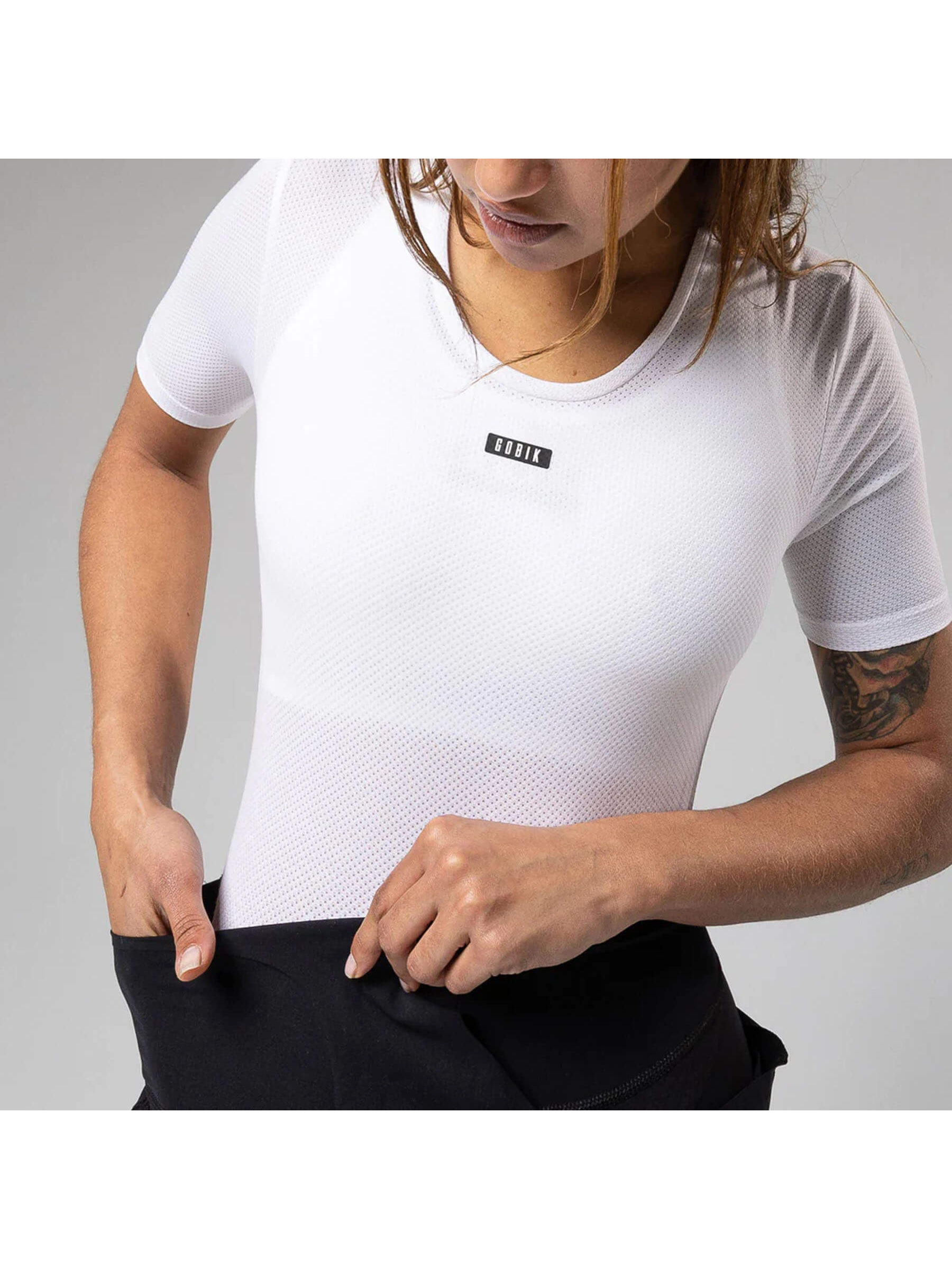 A woman standing in a studio wearing a GOBIK women's short-sleeved base layer in white with a v-neck bib shorts design, ready for a cycling session.